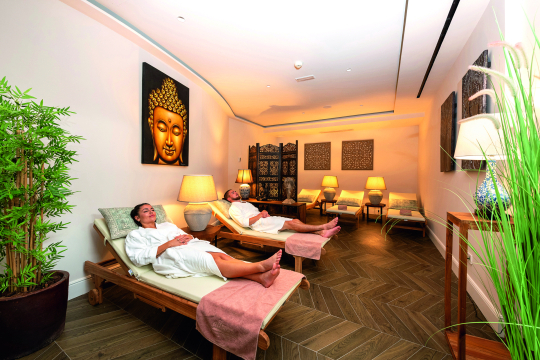 Mann und Frau entspannen im Bademantel auf einer Holzliege in einem Wellnessbereich mit Asiatischer Dekoration an den W&auml;nden